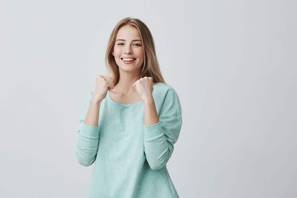 lucky young female employee rejoicing success at work smiling broadly keeping fists clenched beautiful blonde woman in light blue sweater feeling happy and excited posing scaled